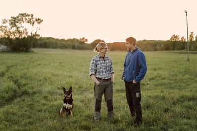 Happy mature couple with dog sitting on field during sunset
