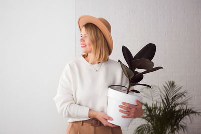 Portrait of young woman wearing hat against wall with big pot ficus home plant