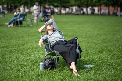 Work-weary woman take break relaxed reclines on chair in open public space on lawn hands over eyes.