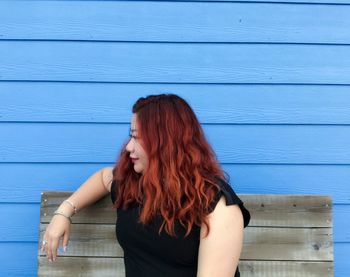 Woman with long hair looking away while sitting on bench against wooden wall