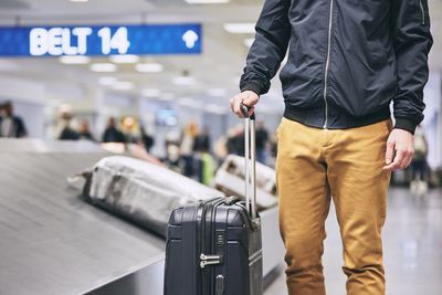 Midsection of young man with luggage standing by conveyor belt at airport