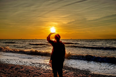 Silhouette woman shielding eyes while standing at beach against orange sky