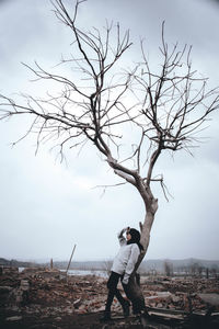 Full length of woman leaning on bare tree against sky
