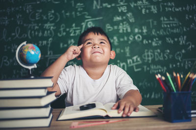 Cute boy studying at table against blackboard