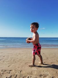 Full length of a baby boy holding coconut on a sandy beach against sky