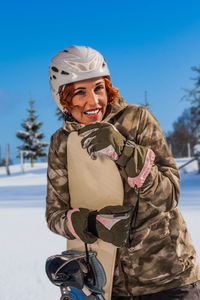 Smiling woman with snowboard looking away while standing on snow covered field against blue sky