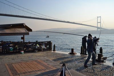 People fishing by river against bosphorus bridge during sunny day