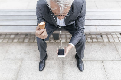 Senior businessman with ice cream cone sitting on bench looking at cell phone