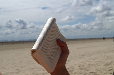 Cropped hand holding book at beach against cloudy sky