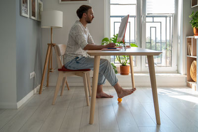 Man freelancer working on computer at home doing recovery foot exercise using massage silicone ball.