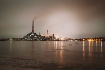 View of illuminated buildings by factory at waterfront against sky