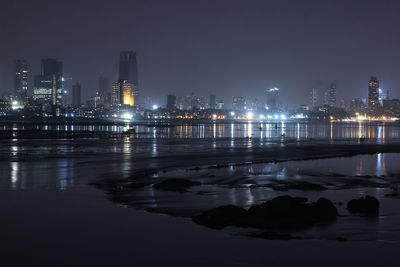 Illuminated buildings by river against sky at night