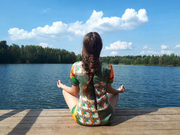 Rear view of woman sitting by lake against sky