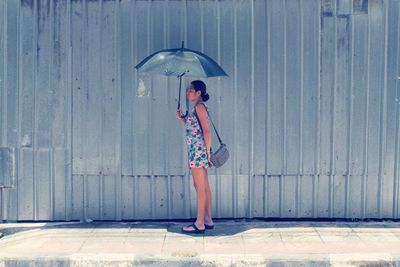 Side view of woman holding umbrella standing against corrugated iron