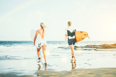 Rear view of friends with surfboards walking towards sea on shore at beach against sky