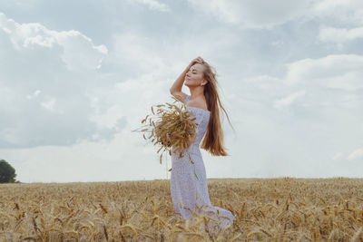 Young blonde woman with long hair stands in a wheat field with an armful of ripe ears of corn.