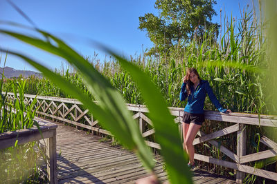 Portrait of young woman standing on footbridge by railing at field during sunny day