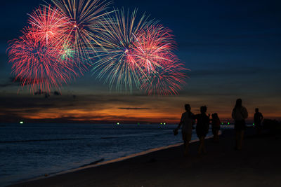 Silhouette people watching firework display at beach against sky at night