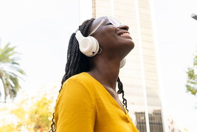 Side view of delighted african american female meloman listening to songs in headphones while standing on street with building in city