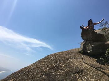 Low angle view of mid adult woman gesturing while sitting on rock against blue sky