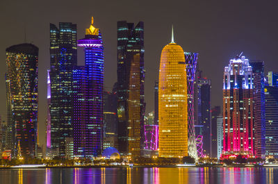 Illuminated buildings by river against sky at night