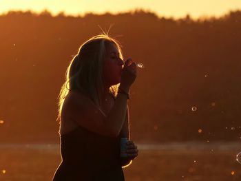 Side view of woman blowing bubbles with wand during sunset