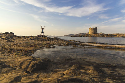 Young woman with raised arms standing on rocks in a beautiful seascape at sunset