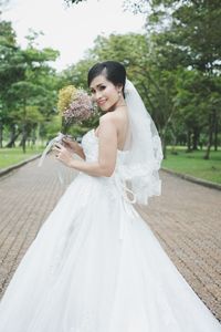 Portrait of smiling bride holding bouquet on footpath at park