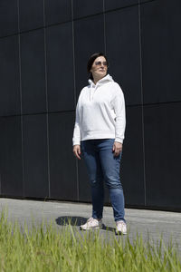 Full length sportive mature brunette woman in white hoody, blue jeans and sunglasses looks away