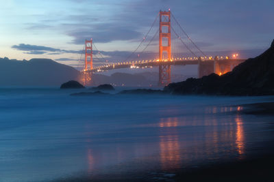 Illuminated suspension bridge over sea against sky