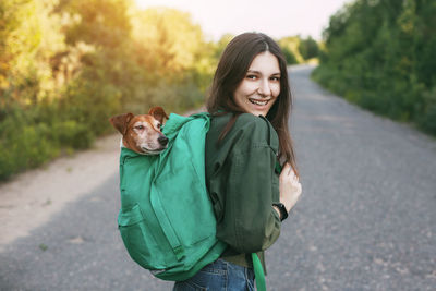 A smiling girl is holding a green backpack on her shoulder, from which a cute dog 