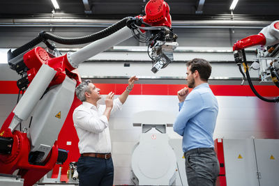 Mature engineer explaining robotic arm to colleague while standing in factory