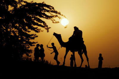 Silhouette people with camel on field against clear orange sky