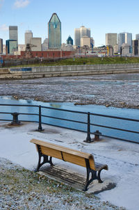 Empty bench by river in city against sky