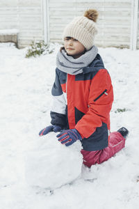 A cute caucasian girl of 9 years old rolls a snowman from the snow in the backyard of the house. 