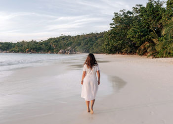 Rear view of woman in white dress walking on tropical sandy beach.