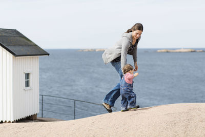 Mother with baby girl walking at beach by sea against clear sky