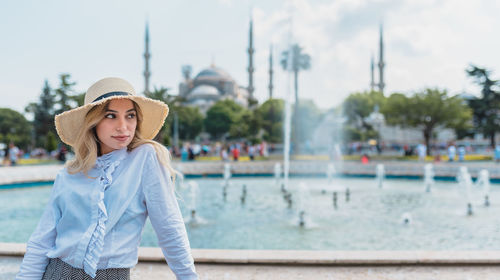 Thoughtful young woman wearing hat while standing against fountain