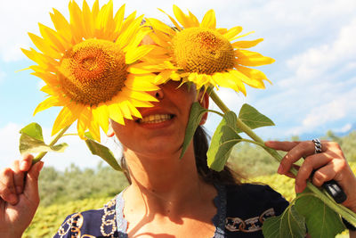 Caucasian brunette girl hides her eyes and part of her face behind two blooming yellow sunflower