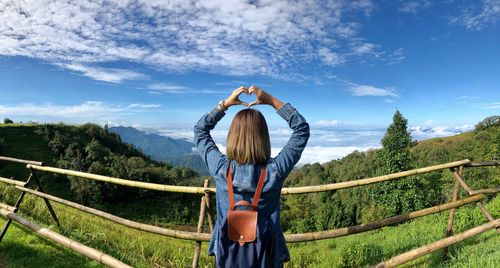 Rear view of woman making heart shape with hands while standing against sky