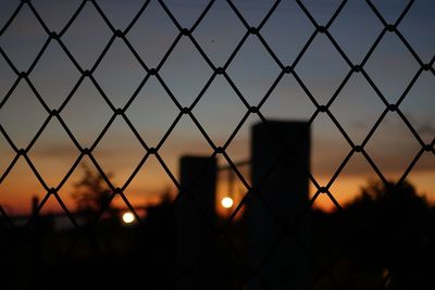 Silhouette man seen through chainlink fence against sky during sunset