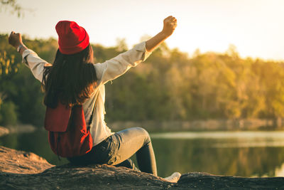 Rear view of woman sitting with arms outstretched by lake