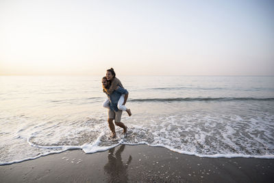 Cheerful man giving piggyback to girlfriend while walking on shore at beach against clear sky during sunset