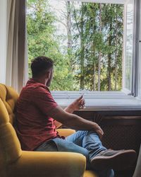 Side view of man having wine in glass while looking through window on chair at home