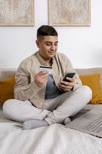 Young man smiling while surfing the web on his smartphone and holding credit card 