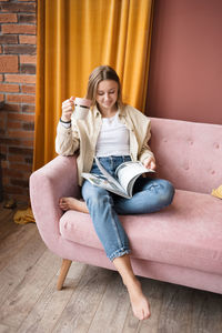 Woman sits on pink sofa in living room with magazine and cup of tea.