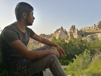 Side view of young man sitting on rock against sky