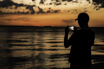 Silhouette man photographing sea with mobile phone during sunset