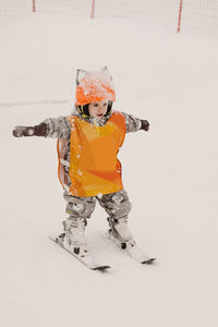 Full body of adorable little child in warm winter overalls and protective helmet standing on skis while practicing skiing on snowy terrain