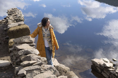 High angle view of young woman standing on rock against lake with reflection of sky